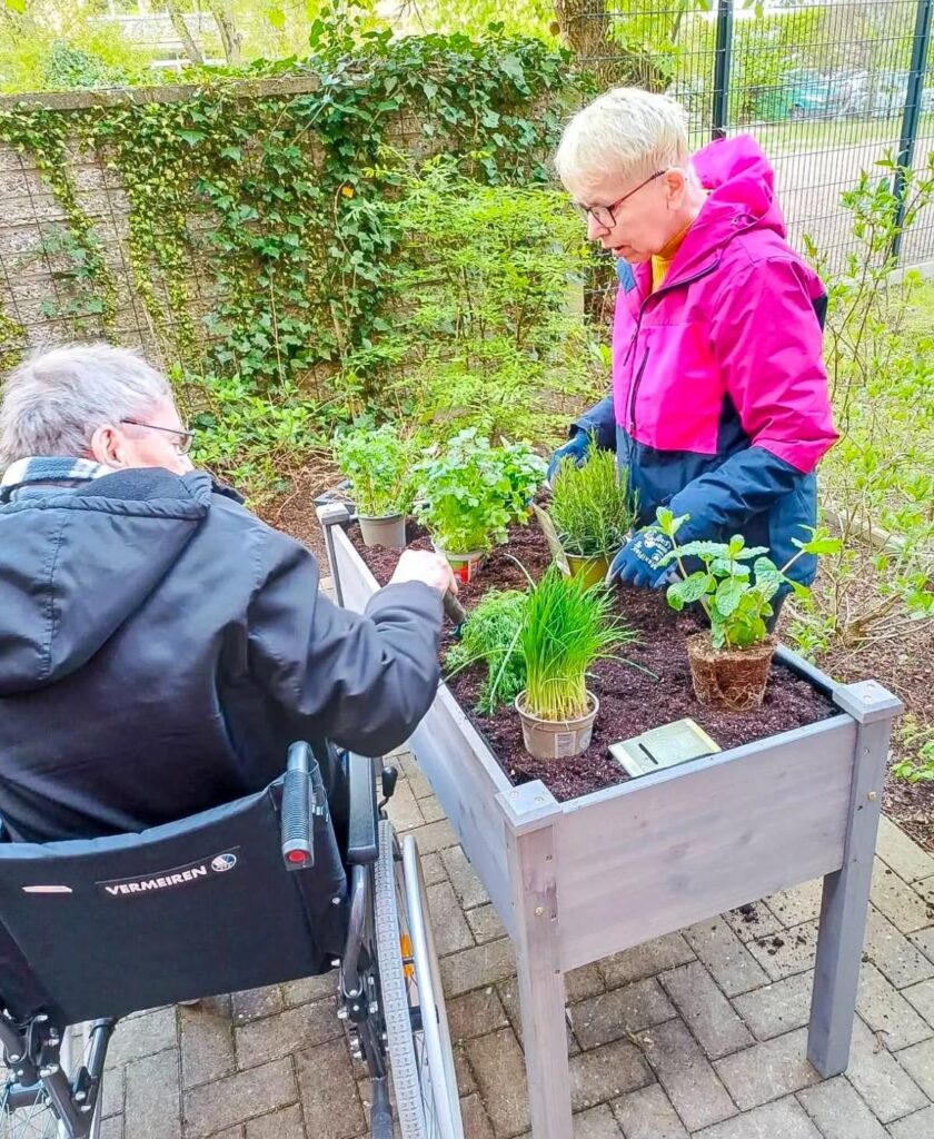 Kunde einer Intensivpflege-WG arbeitet gemeinsam mit einer Fachkraft an einem Hochbeet im Garten